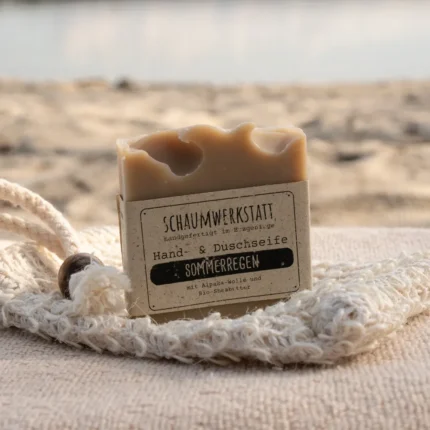 Braune Alpakaseife Sommerregen mit einer Banderole aus Graspapier am Strand. Im Hintergrund ist Wasser zu sehen.