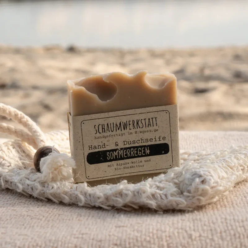 Braune Alpakaseife Sommerregen mit einer Banderole aus Graspapier am Strand. Im Hintergrund ist Wasser zu sehen.