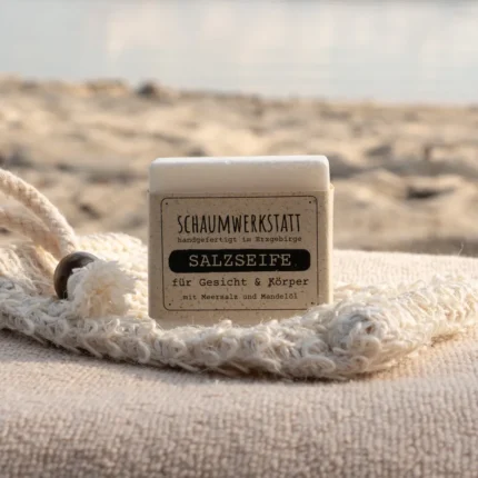Salzseife mit einer Banderole aus Graspapier arrangiert auf einem Sisalsäckchen und einem Badetuch am Strand