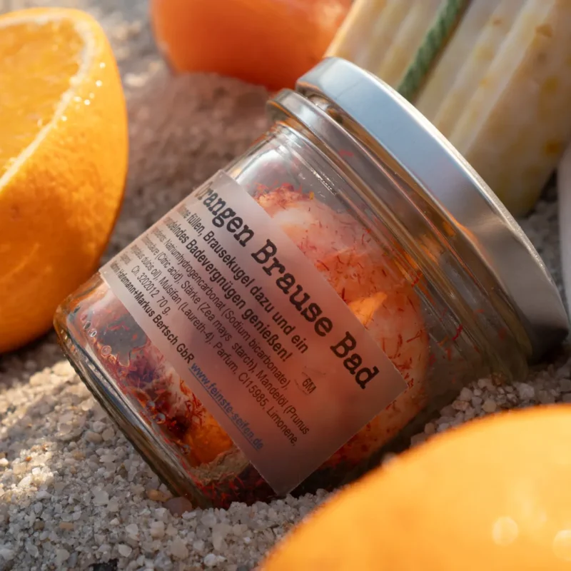 Badekugel Orange im Glas mit Orangenscheiben im Sand arrangiert
