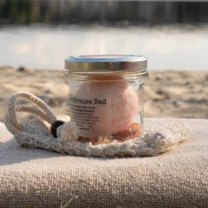 Badekugel Rose im Glas auf Sisalsäckchen und Badetuch am Wasser mit natürlicher, ruhiger Umgebung