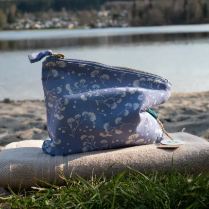 Blaue Kosmetiktasche aus Stoff mit weißen Blüten am Strand arrangiert